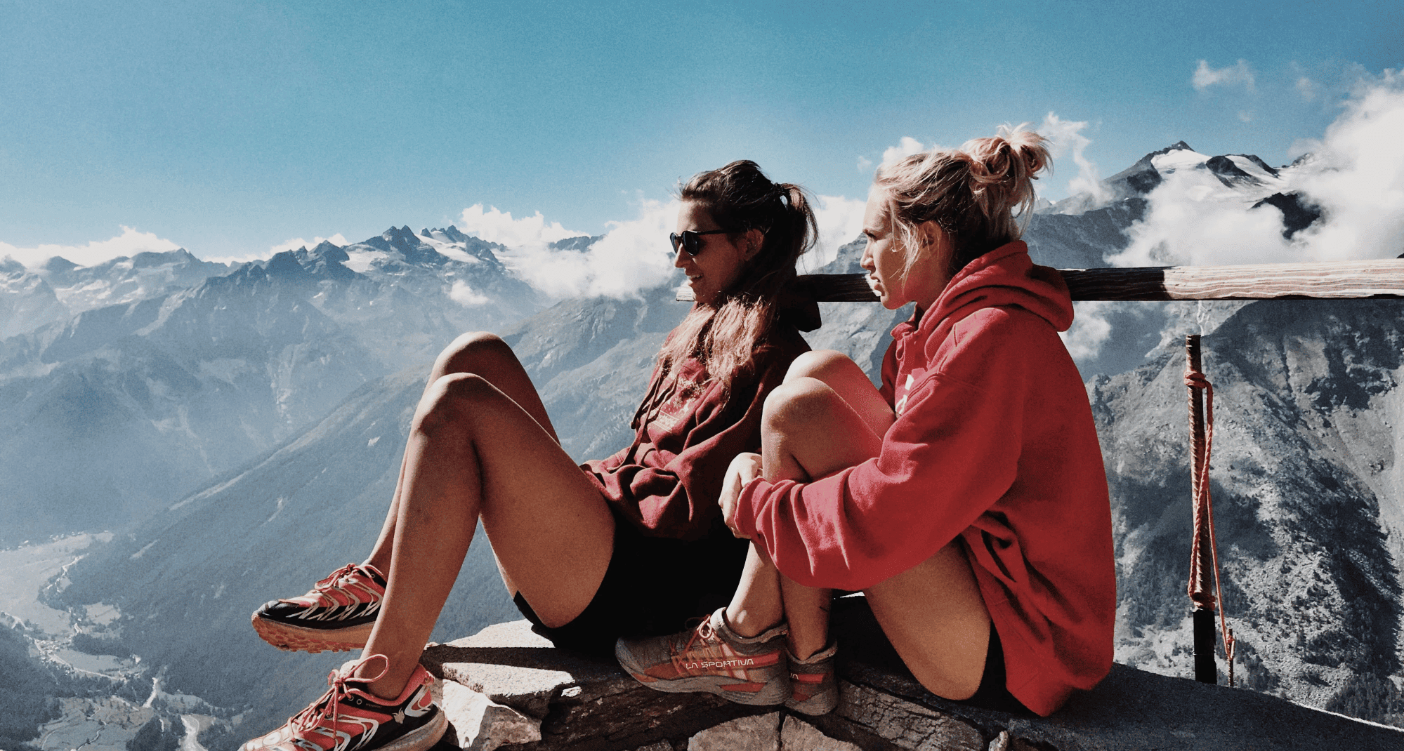 Two women sit on the edge of a mountain summit, and look out over the view.