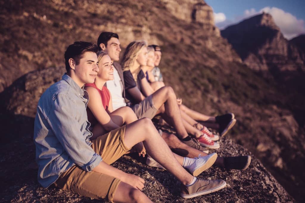 Group of teens sit in a row on a mountain and watch the sunset