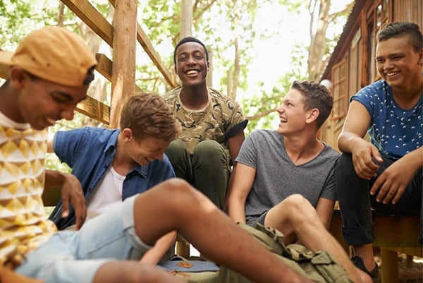 Group of teen males laugh while sitting on a wooden deck